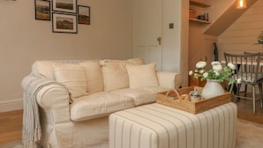 A living room with a beige sofa, a striped ottoman, and a basket on the ottoman. There are framed pictures on the wall and a wooden staircase in the background. - Stag Cottage (Minehead)