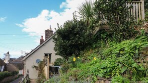 A quaint house with a stone wall and a wooden gate, surrounded by lush greenery and a clear sky. - Stag Cottage (Minehead)