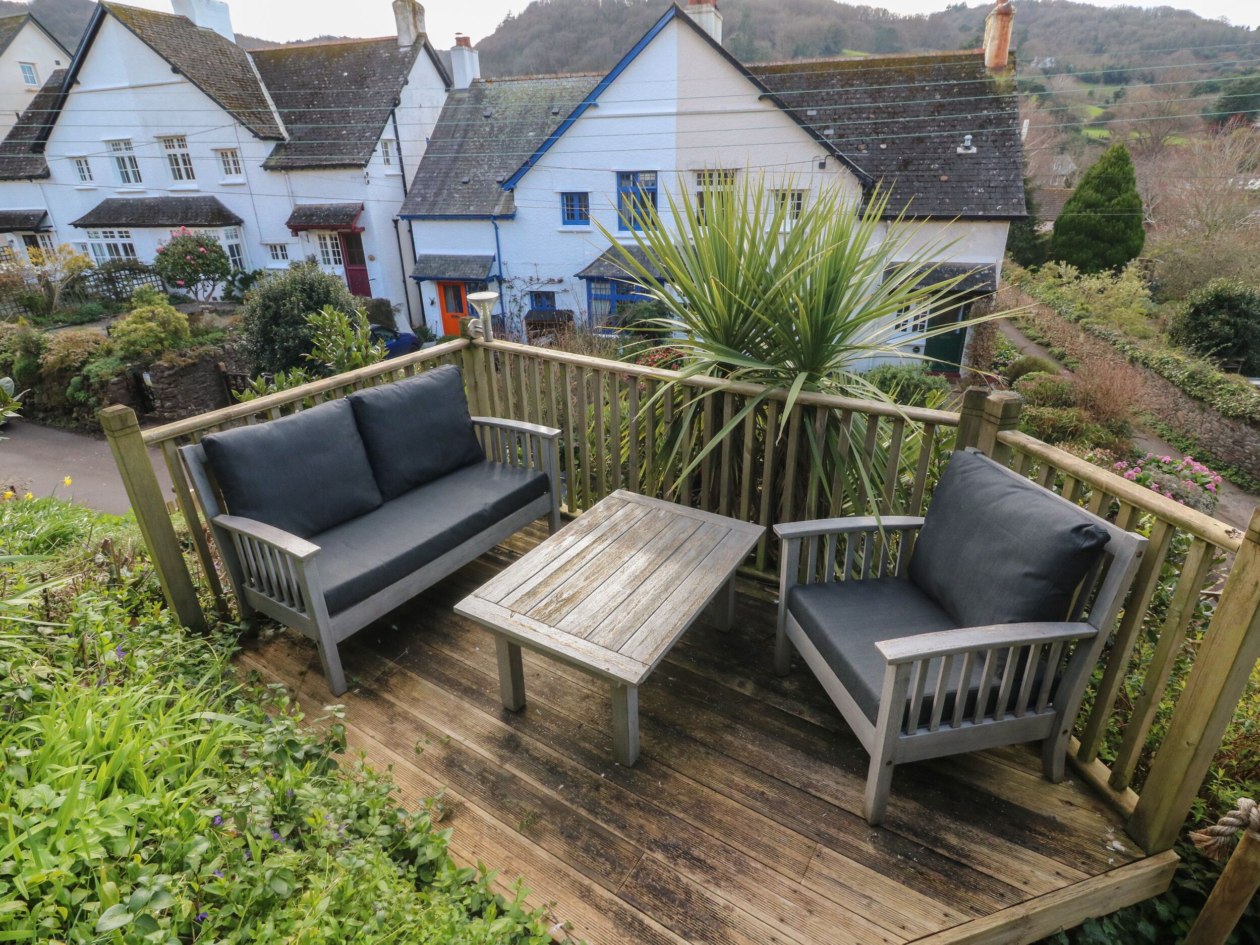 A wooden deck with a sofa and chairs, surrounded by greenery and overlooking a white building with blue accents.