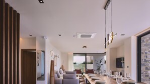 A modern dining area with a long table set for six, a sofa, and a large window offering a view of the outdoors. - Walking distance to Beach, Heated Pool & Rooftop Hot Tub! (Rethymno)