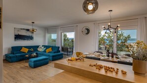 A modern living room with a blue sofa set, a wooden coffee table with food, and a view of the sea through large windows. - Seaview villa Deep Green in Maslenica (Maslenica)