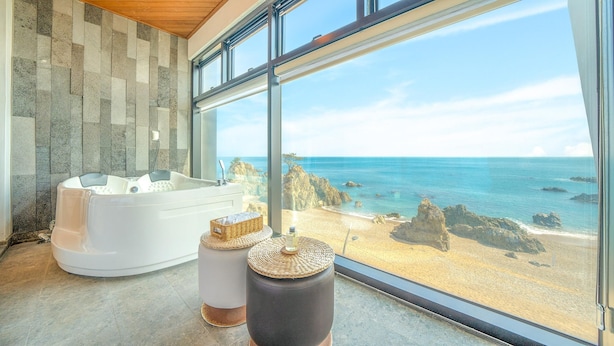 A modern bathroom with a large bathtub, a small stool, and a view of the ocean through large windows. - Gyeongju Blue21 Poolvilla Pension (Gyeongju)