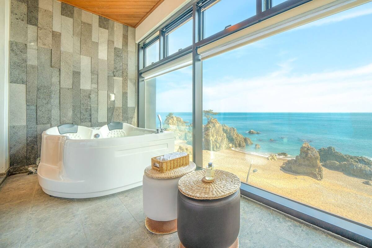 Ein modernes Badezimmer mit einer großen Badewanne, einem kleinen Hocker und Meerblick durch große Fenster.