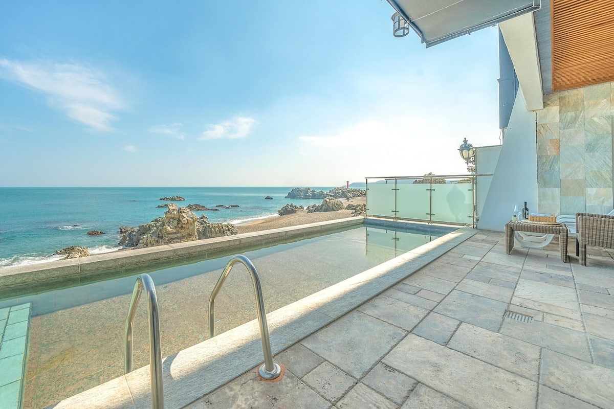 Ein Swimmingpool mit Meerblick, eine Lounge mit Tisch und Stühlen sowie eine deckenartige Terrasse mit Steinplatten.