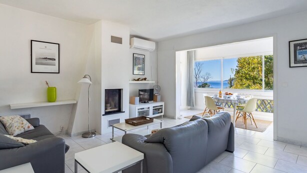 Un salon moderne avec un canapé gris, une table basse blanche et un téléviseur posé sur un meuble blanc. Il y a un balcon avec une table et des chaises, et une vue sur la mer.