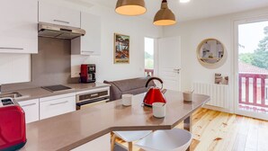 Eine moderne Küche mit weißen Schränken, einem eingebauten Backofen, einer Kaffeemaschine und einem roten Wasserkocher auf der Kücheninsel. Zwei Pendelleuchten hängen über der Insel.