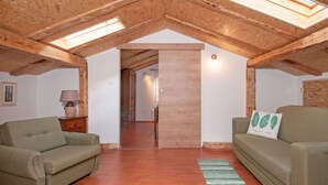 Quarto no sótão com vigas de madeira, um claraboia, dois sofás verdes, uma porta de madeira e um tapete verde listrado.