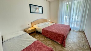 Habitación de hotel con dos camas, una mesita de noche, una lámpara de noche, un cuadro en la pared y una ventana con cortinas translúcidas.