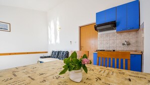 A room with a bed, a table with a coffee-themed tablecloth, a potted plant, and a kitchenette with blue cabinets.
