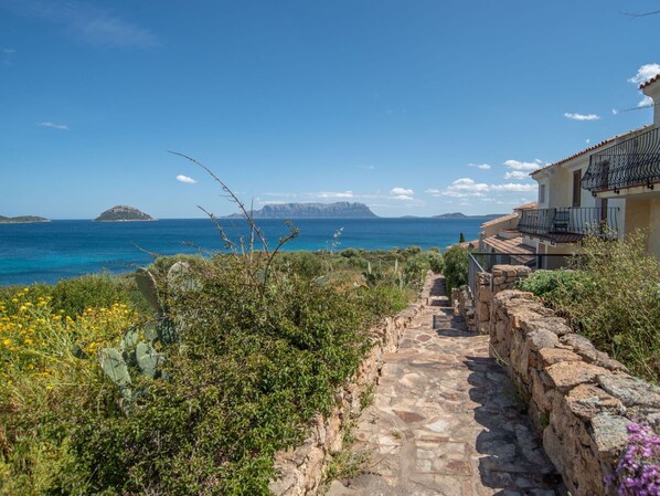 A stone pathway leading to a building with a balcony overlooking a coastal view.