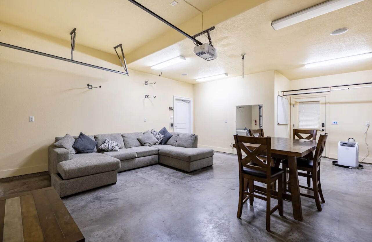A spacious living area with a gray sectional sofa, a wooden dining table with chairs, and a ceiling-mounted projector.