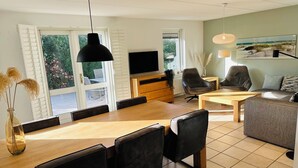 Un salón moderno con una mesa de comedor de madera, sillas de cuero negro, un televisor sobre un mueble de madera y un ventanal con contraventanas.