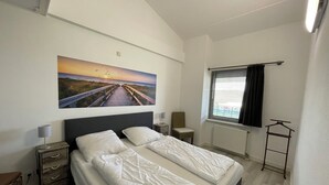 Un dormitorio con una cama grande, una mesita de noche, una silla, una ventana con persianas y un cuadro colgado en la pared que muestra un atardecer sobre un puente.