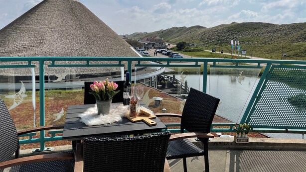 Una mesa con un florero, una botella de vino y copas, con vistas a un cuerpo de agua y un edificio con techo de paja.