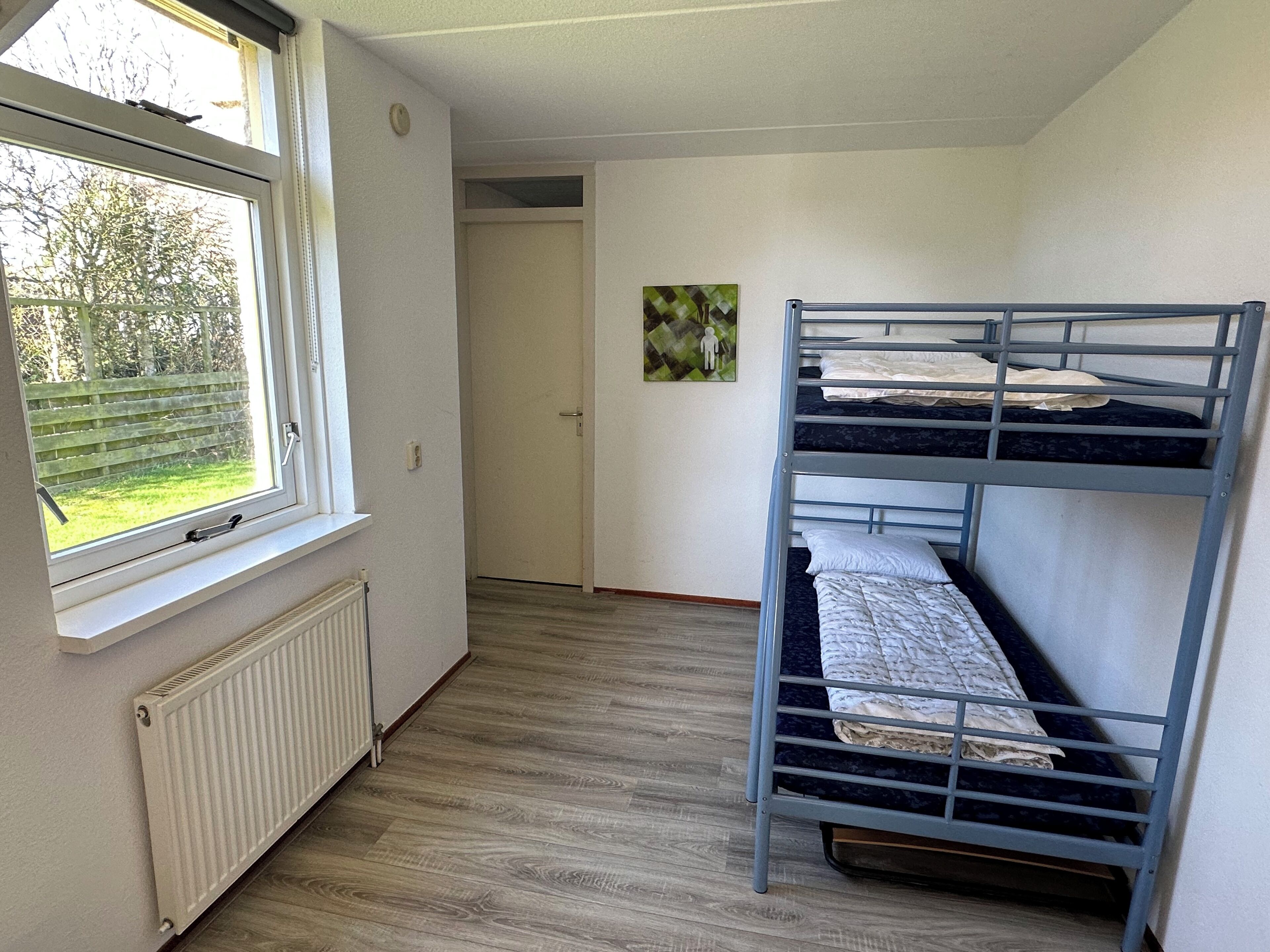 A dormitory room with bunk beds, a radiator, a window with blinds, and a door leading to another room.