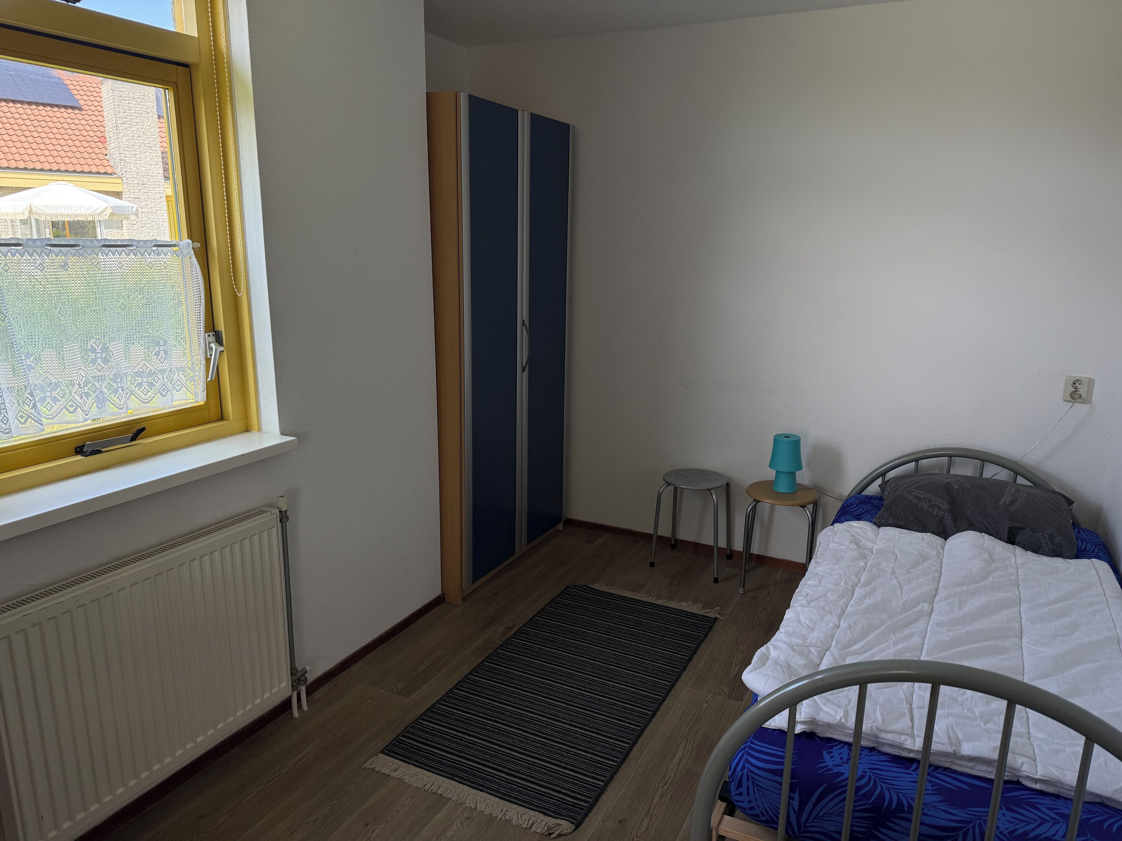 Un dormitorio con cama, radiador, ventana, un taburete pequeño y armario.