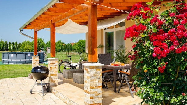 Eine Terrasse im Freien mit einer Holz-Pergola, Steinpfeilern, Korbmöbeln und einem Grill.