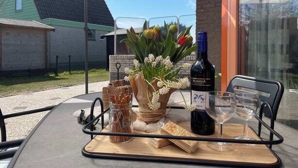 A table set outdoors with a bottle of wine, glasses, a jar of pretzels, a basket of flowers, and a sign indicating a restaurant. - 3 bedroom accommodation in Julianadorp aan Zee (Julianadorp aan Zee)