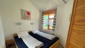 A bedroom with two beds, a window with curtains, a small table, and a wall-mounted lamp. - 3 bedroom accommodation in Julianadorp aan Zee (Julianadorp aan Zee)