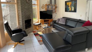 A living room with a fireplace, black leather sofa, television, bookshelf, and a large window.