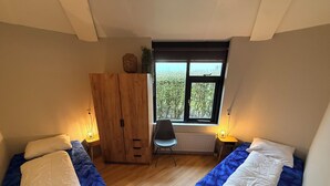 A bedroom with two beds, a wooden wardrobe, a chair, a window with a view, and two bedside lamps.