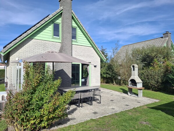 Een huis met een groen dak, een schoorsteen, een terras met een tafel en stoelen, en een stenen buitenoven.