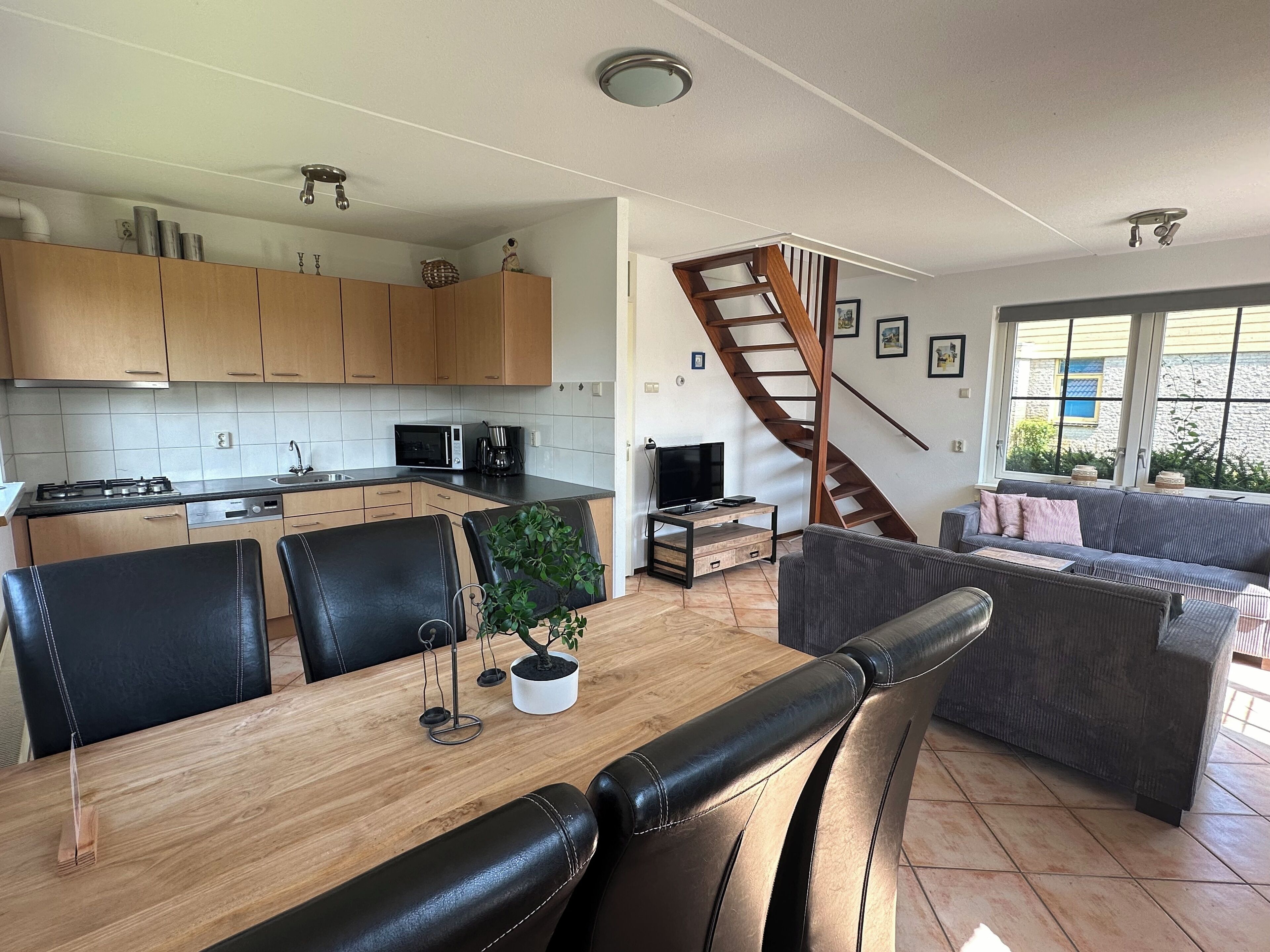 Een moderne keuken met houten kasten, een eethoek met een houten tafel en zwarte leren stoelen, een grijze bank en een houten trap.