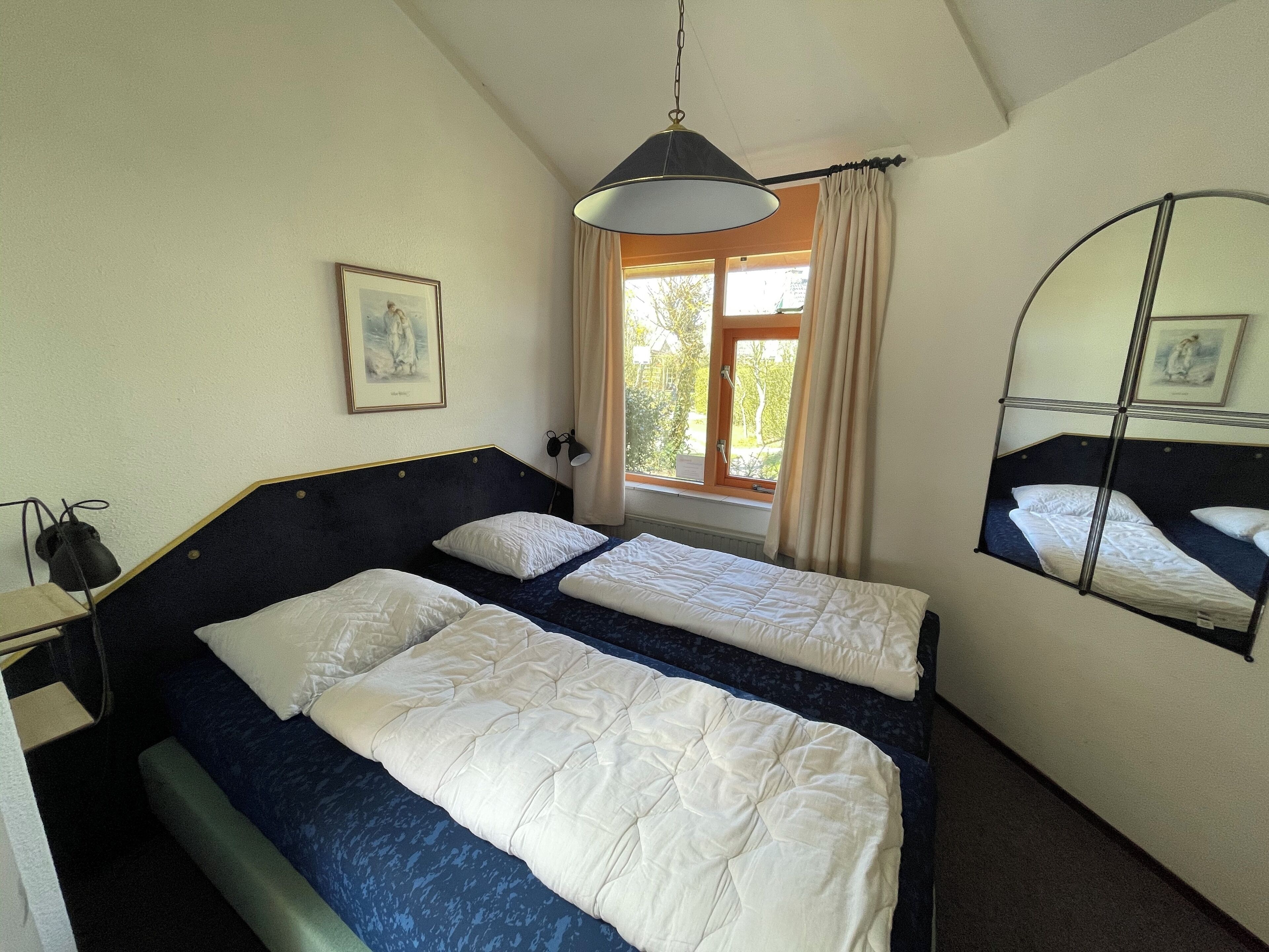 Een slaapkamer met twee bedden, een raam met gordijnen, een hanglamp en een ingelijst kunstwerk aan de muur.