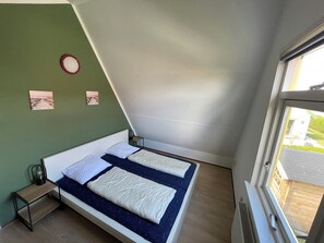 A small, modern bedroom with a sloped ceiling, two beds with white and blue bedding, a small table with a lamp, and a window with a view of the outside.