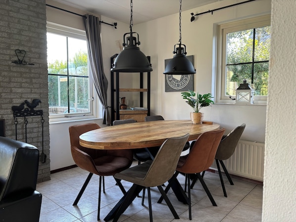A dining area with a wooden table and chairs, a hanging light fixture, and a view of greenery outside.