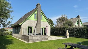 Una casa de dos pisos con techo verde y fachada de ladrillo, un patio con mobiliario exterior y un jardín.
