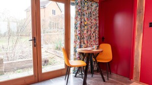 A room with a sliding glass door leading to a garden, a red door, and a table with two orange chairs. - Jossies Snug - peaceful, private garden/hot tub, swimming lake, Spa and Wellness (Bletchingley)