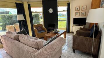 Een woonkamer met een bank, fauteuils, een salontafel, een open haard, een klok, een televisie en ingelijste foto's aan de muur.