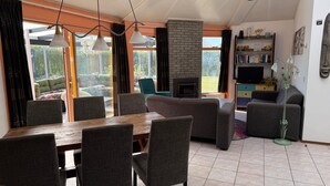 A dining area with a wooden table and grey upholstered chairs, a fireplace, and a bookshelf with various items. - 3 bedroom accommodation in Julianadorp aan Zee (Julianadorp aan Zee)
