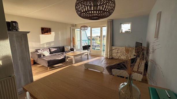 Ein modernes Wohnzimmer mit einer Couch, einem Couchtisch und einem Essbereich mit einem Holztisch. Ein großes Fenster bietet einen Blick ins Freie.