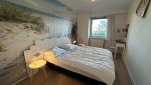 Ein Schlafzimmer mit einem großen Bett, einem kleinen Nachttisch, einem Sessel, einem Schreibtisch und einem Fenster mit Vorhängen.