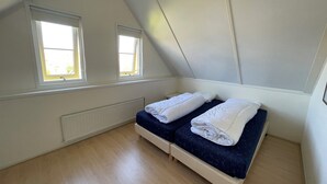 Pieni ullakkohuone, jossa on yksi huone, kaksi ikkunaa, patteri ja sänky, jossa on valkoiset, siististi taitetut lakanat.