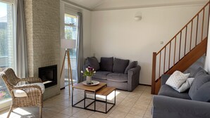 Een woonkamer met een grijze bank, een rieten stoel, een klein houten tafeltje met een vaas, een open haard en een trap.