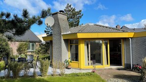 Una casa moderna con fachada amarilla, una chimenea de piedra y un gran ventanal que da a un porche cubierto.