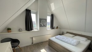 A bedroom with a sloped ceiling, a bed with white bedding, a chair, a radiator, and a window with curtains.