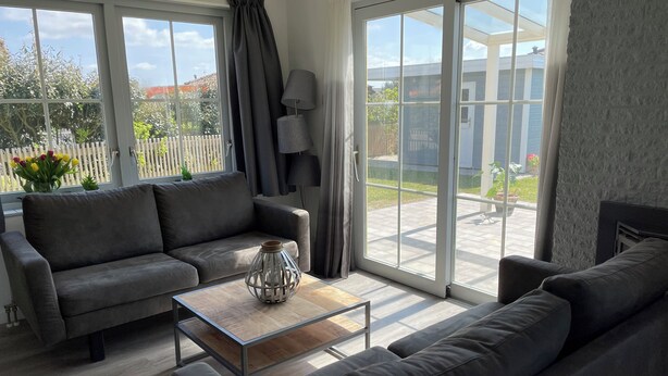 A modern living room with a grey sofa, a coffee table, and a lamp. There are large windows with curtains and a view of the outside.