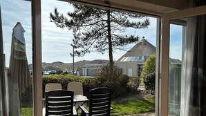 A view from a room with a large window, featuring a patio with a table and chairs, a tree, and a building in the background. - 2 bedroom accommodation in Julianadorp aan Zee (Julianadorp aan Zee)