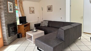 Een woonkamer met een hoekbank, een open haard, een televisie en een bijzettafeltje met een schaal.