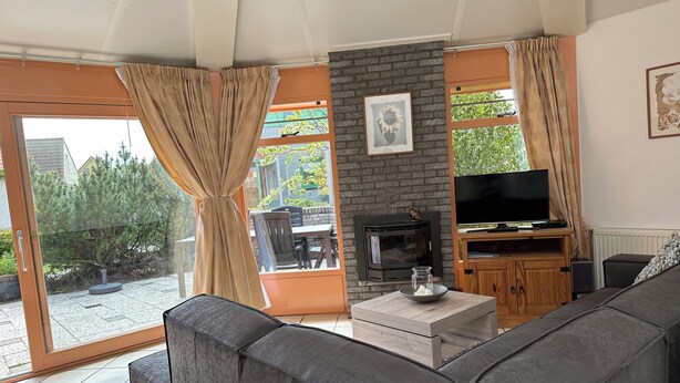 Een woonkamer met een grijze bank, een open haard, een televisie en uitzicht op groen.
