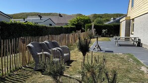 Een tuin met rieten stoelen, een houten hek en een terras met betegelde vloer en picknicktafels.