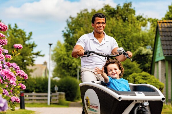 A man riding a bicycle with a child in a child seat, in a park with greenery and a building in the background.
