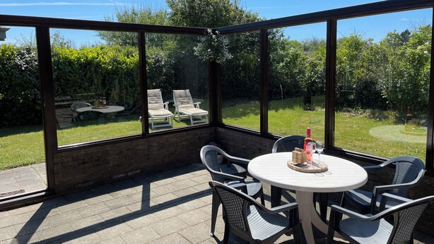 Un patio coperto con un tavolo rotondo e sedie, vista su un giardino con sdraio e una bottiglia di vino sul tavolo.