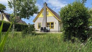 Une maison à l’extérieur jaune et gris, avec une cheminée et un petit étang où nagent des canards.