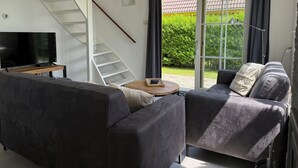 Un salon moderne avec un canapé gris, une table basse, un téléviseur posé sur un meuble en bois, un escalier et une porte-fenêtre coulissante donnant sur un jardin.
