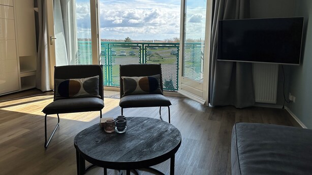 Een moderne woonkamer met een groot raam, twee fauteuils, een ronde tafel en een flatscreen televisie.